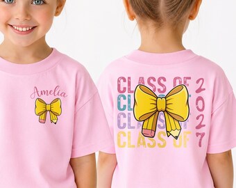 May include: A light pink short-sleeve t-shirt featuring a yellow bow-shaped pencil graphic. The front displays the name Amelia above the bow. The back features the text Class of 2027 in a stacked, multi-colored font behind a large yellow pencil bow.