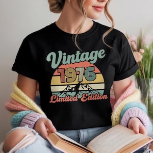 May include: Black t-shirt with a retro design featuring the words "Vintage 1976" above a colorful sunset graphic and mountain range. The text "Limited Edition" is below the graphic. The shirt is a classic crew neck.