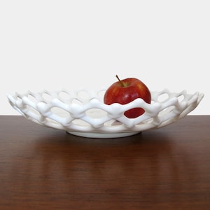 Könnte beinhalten: Weiße Keramikschale mit Gitterdesign. Die Schale ist flach und hat einen erhöhten Rand. Ein roter Apfel liegt in der Mitte der Schale.