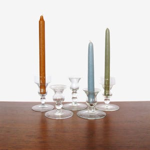 May include: Four glass candle holders with three candles. The candles are brown, blue, and green. The candle holders are clear glass with a flared base and a small cup on top.
