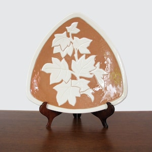 May include: A triangular ceramic plate with a white rim and a brown center. The plate features a raised white leaf design. The plate is displayed on a dark brown wooden stand.