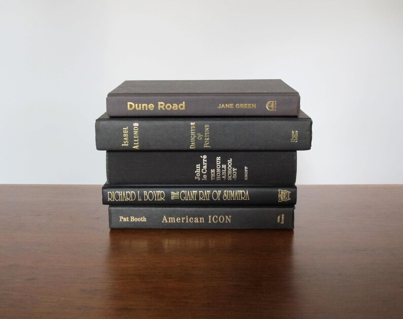 May include: A stack of six black hardback books with gold lettering on the spines. The titles are "Dune Road", "Daughter of Fortune", "The Honour School", "The Giant Rat of Sumatra", and "American Icon".