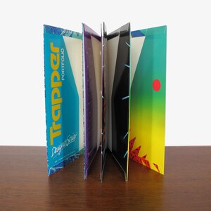 May include: A Trapper Portfolio folder standing open, displaying multiple interior panels with vibrant graphic designs. The left panel features the word Trapper in yellow text on a blue background, while other sections show black, purple, and yellow-to-green gradients with abstract patterns.