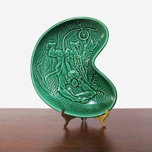 May include: A green ceramic wall plate with a raised design of a monkey hanging from a vine. The plate is shaped like a crescent moon and has a gold metal stand.