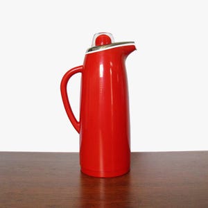 May include: A red thermos with a white lid and a silver spout. The thermos is sitting on a wooden table.