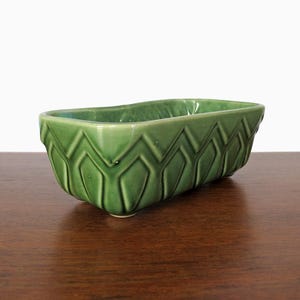 May include: A rectangular, green ceramic planter with a geometric pattern. The planter has a glossy finish and is elevated on small feet. The design features a repeating zig-zag pattern around the sides, adding a decorative touch to this vintage-style container.