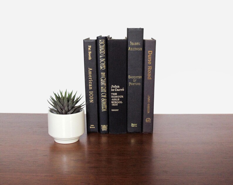 May include: A collection of six black hardback books with gold lettering on a dark brown wooden surface. The books are arranged vertically, with the tallest book in the centre. The titles of the books are "Dune Road", "Daughter of Fortune", "The Honour Able School-Boy", "Giant Rat of Sumatra", "American Icon", and "The Giant Rat of Sumatra".