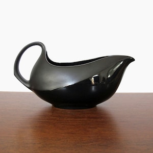 May include: A black ceramic gravy boat with a curved handle. The boat has a glossy finish and a smooth, rounded shape.