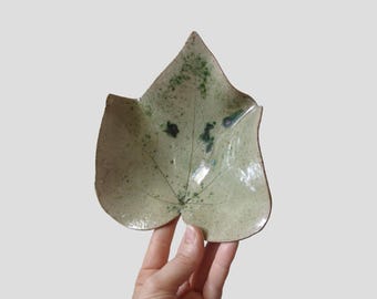 May include: A handmade ceramic leaf-shaped dish. The dish is a pale green color with darker green speckles and veining details. The edges are slightly raised, creating a shallow bowl. A unique piece for serving or display.