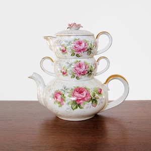 May include: Stacked porcelain teacups and teapot set. The set features a white background with pink roses and green leaves. The teapot has a gold handle and spout. The lid is topped with a pink rose.