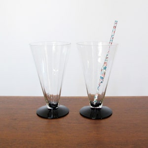 Könnte beinhalten: Zwei klare Glas-Trinkgläser mit schwarzen Füßen. Die Gläser sind hoch und schmal, mit einem ausgestellten oberen Rand. Ein gemusterter Strohhalm befindet sich in einem der Gläser.