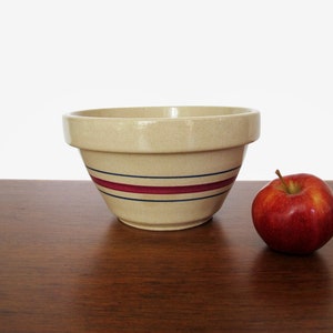 May include: A cream-colored ceramic mixing bowl with three red and blue stripes around the rim. The bowl is sitting on a wooden surface next to a red apple.