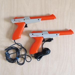 May include: Two orange and gray Nintendo Zapper light guns with black cords. The guns are designed to look like pistols and are a classic video game accessory.