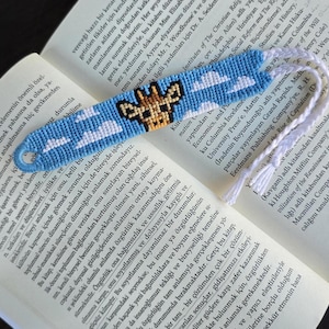 May include: Handmade bookmark featuring a giraffe design with a blue sky and cloud pattern. The bookmark is made of woven threads and has white tassels. It is resting on the pages of a book.