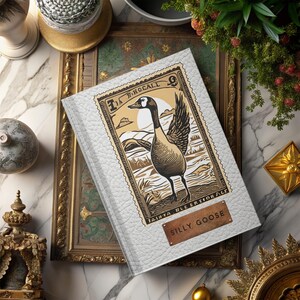 Pode incluir: Um diário encadernado em couro branco com uma ilustração de estilo vintage de um ganso em um selo postal. O diário tem uma placa de título dourada que diz "Silly Goose".