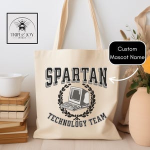 Może przedstawiać: Kremowa torba typu tote z napisem "SPARTAN TECHNOLOGY TEAM" w kolorze czarnym i szarym, z grafiką komputera. Torba ma długie uchwyty i logo w lewym górnym rogu. Można dodać niestandardową nazwę maskotki.