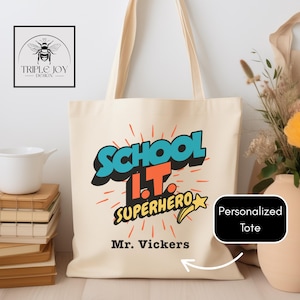 Może przedstawiać: Kremowa torba typu tote z napisem "SCHOOL I.T. SUPERHERO" w stylu komiksowym. Imię "Mr. Vickers" jest wydrukowane poniżej. Torba ma długie uchwyty i logo w lewym górnym rogu.