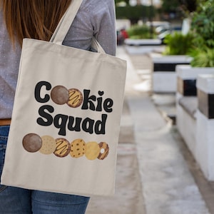 May include: A beige tote bag with the words "Cookie Squad" in black, with a heart symbol. Below the text are watercolor illustrations of various cookies. The bag is carried over the shoulder.