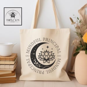 May include: Cream-colored tote bag with the words "Mindful Principal" encircling a crescent moon and lotus flower design. The bag has long handles and a rectangular shape. A logo with a bee is visible in the upper left corner.