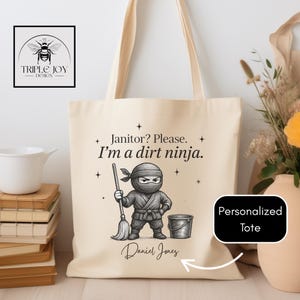 Peut inclure: Sac fourre-tout crème avec un motif de ninja et le texte "Janitor? Please. I'm a dirt ninja." Le motif comprend un ninja avec un balai et un seau. Les mots "Personalized Tote" sont également visibles.