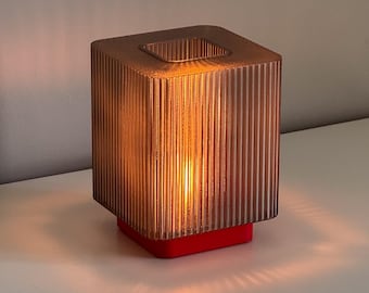 May include: A modern, cube-shaped lamp with a ribbed, bronze-colored shade. The lamp sits on a red base, and the warm light from within casts shadows. The lamp is a decorative lighting fixture.