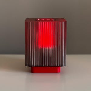 May include: A red and clear, ribbed, rectangular lamp with a bright red interior glow. The lamp sits on a red base and has a square opening at the top. The lamp is on a white surface with a gray background.