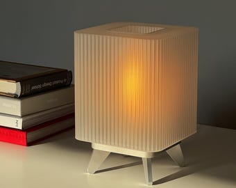 May include: A modern, square table lamp with a ribbed, translucent shade. The lamp has a warm, glowing interior and sits on a white base with three angled legs. The lamp is on a white surface with books in the background.