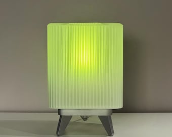 May include: A modern, illuminated table lamp with a ribbed, lime green shade. The lamp has a rectangular shape and sits on a silver base with three angled legs. The light glows from within, creating a soft, ambient glow.