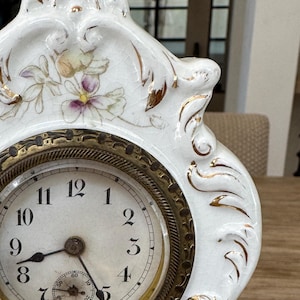 Puede incluir: Reloj de chimenea de porcelana blanca con detalles dorados y diseños florales. La esfera del reloj tiene agujas negras y números romanos.