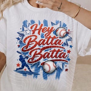 May include: White t-shirt with the text "Hey Batta Batta" in red and blue script, along with baseball graphics. The design includes stars, baseballs, and a baseball player silhouette. The shirt has rolled-up sleeves.