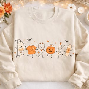 May include: A cream-colored crewneck sweatshirt featuring a graphic print of smiling medical-themed characters holding hands. The design includes an IV bag, scrubs, a stethoscope, a jack-o'-lantern, a thermometer, and a bandage, with small bats and hearts.