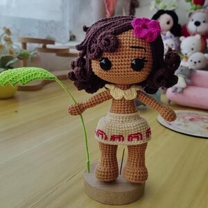 May include: Crocheted doll with brown skin, curly brown hair, and a pink flower. The doll wears a beige skirt with red trim and holds a green leaf. It stands on a wooden base. The doll is approximately 6 inches tall.