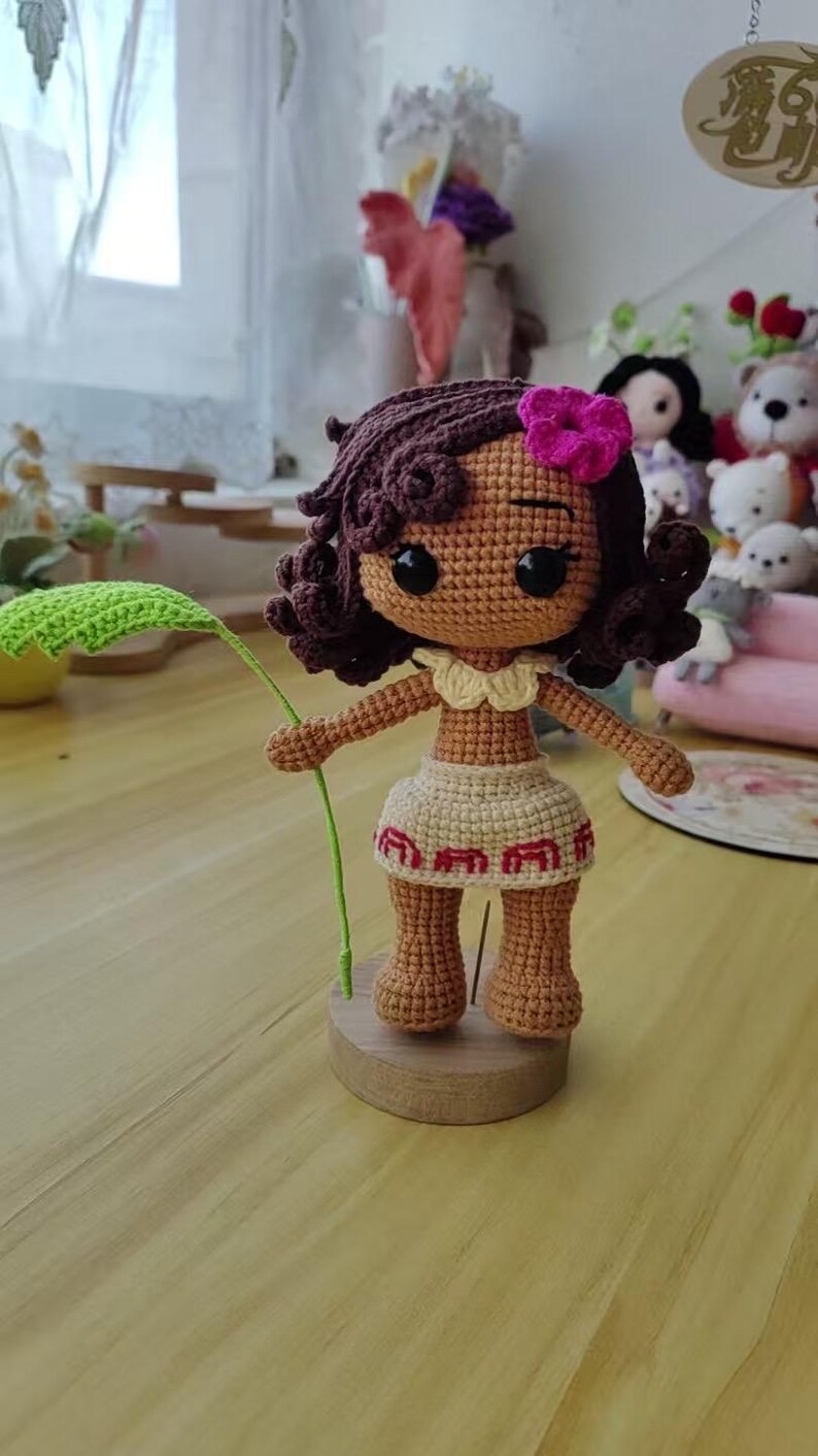May include: Crocheted doll with brown skin, curly brown hair, and a pink flower. The doll wears a beige skirt with red trim and holds a green leaf. It stands on a wooden base. The doll is approximately 6 inches tall.