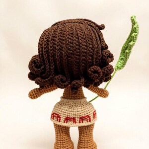 May include: A handmade crochet doll with brown hair and skin, wearing a beige skirt with red accents. The doll is holding a green leaf. The doll's hair is styled in curls. The doll is viewed from the back.