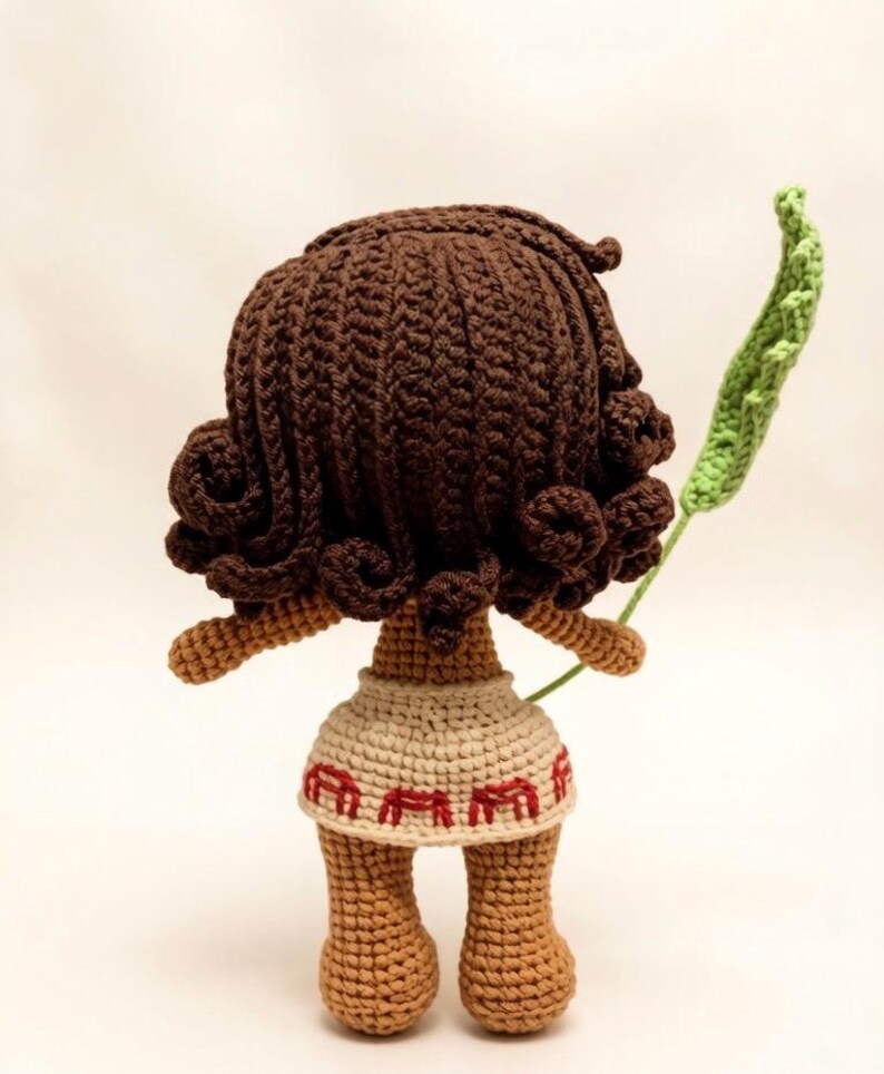 May include: A handmade crochet doll with brown hair and skin, wearing a beige skirt with red accents. The doll is holding a green leaf. The doll's hair is styled in curls. The doll is viewed from the back.