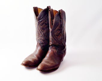 Può includere: Un paio di stivali da cowboy in pelle marrone con cuciture decorative. Gli stivali hanno la punta affusolata e un tacco impilato. La pelle è di un ricco colore marrone scuro con cuciture marrone chiaro. Gli stivali sono su sfondo bianco.