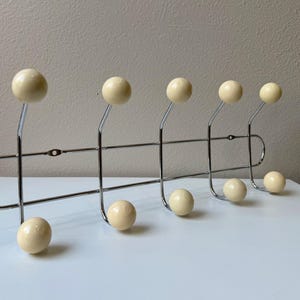 Puede incluir: Un perchero de metal cromado con ocho ganchos en forma de bola de color crema. El perchero tiene una barra horizontal con extremos curvos y soportes verticales para los ganchos. Los ganchos están espaciados uniformemente.