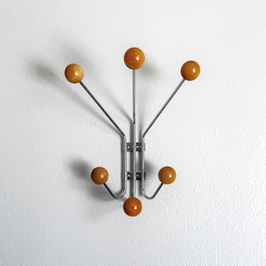 May include: A mid-century style coat rack featuring six wooden ball knobs in a warm, honey colour. The knobs are attached to silver metal rods and a central mounting bracket. The rack is mounted on a white wall.
