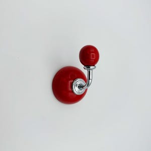 May include: A red ceramic hook with a chrome metal hook arm. The hook is mounted on a white wall.