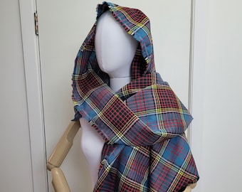 May include: A hooded scarf in a blue, red, yellow, and black plaid pattern. The scarf is draped over a mannequin, showcasing the fringed edges and the fabric's texture. The hood is up, and the scarf is wrapped around the mannequin's neck and shoulders.