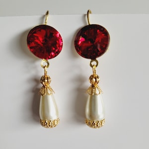 Op de afbeelding: Een paar oorhangers met een rode kristal en een witte parel. De oorhangers hebben een gouden afwerking.