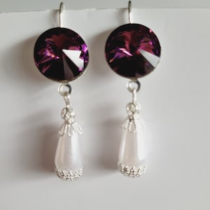 Op de afbeelding: Een paar oorhangers met een paarse kristallen steen en een witte parel. De oorhangers hebben een zilveren afwerking.