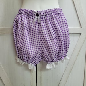 May include: Purple and white gingham bloomers with a white lace trim. The elastic waistband is accented with a small white bow and a dark purple button. These shorts are designed with a gathered, rounded shape.