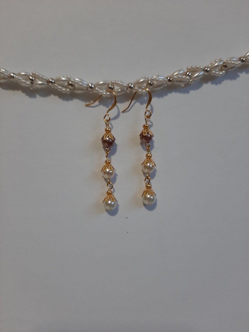 May include: A pair of gold dangle earrings with three white pearls and a brown pearl on each earring.
