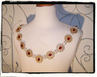May include: A gold-toned necklace with red gemstone accents and faux pearls, displayed on a white mannequin. The necklace features ornate, circular designs with a central red stone, connected by small pearls. The necklace is designed to be worn across the shoulders.