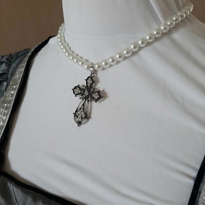 May include: A white pearl necklace with a silver cross pendant. The cross has a detailed design with small stones.