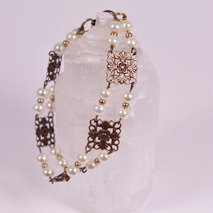 Op de afbeelding: Een delicate armband met een dubbele rij witte parels en filigraan accenten van brons. De armband wordt getoond op een wit selenietkristal.