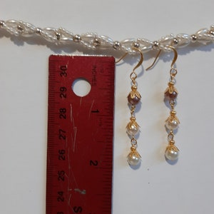 May include: A pair of gold dangle earrings with three white pearls and a brown bead on each earring. The earrings are measured against a ruler.