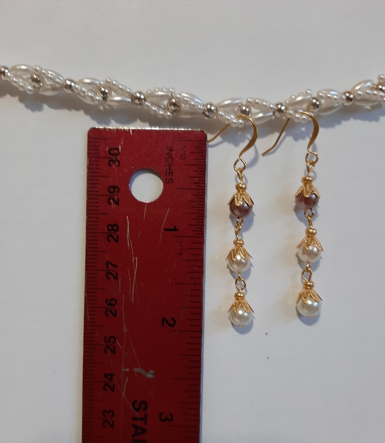 May include: A pair of gold dangle earrings with three white pearls and a brown bead on each earring. The earrings are measured against a ruler.