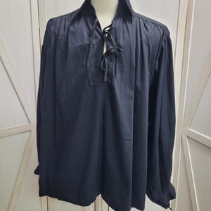 May include: A dark navy blue long-sleeved shirt featuring a pointed collar and a front placket with a lace-up closure using matching fabric ties. The shirt has gathered fabric at the shoulders and cuffs, creating a loose, flowing fit.